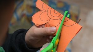 A closeup of a pair of green scissors cutting an orange piece of paper into the shape of a flower